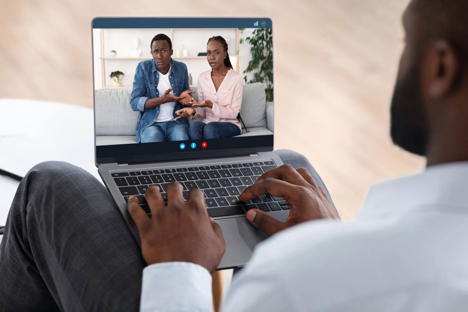 A person engages in online couples therapy via video call, with a couple on a laptop screen discussing marriage counselling, seated on a couch and gesturing. The individual holding the laptop sits cross-legged indoors. Rosen Couples Counselling