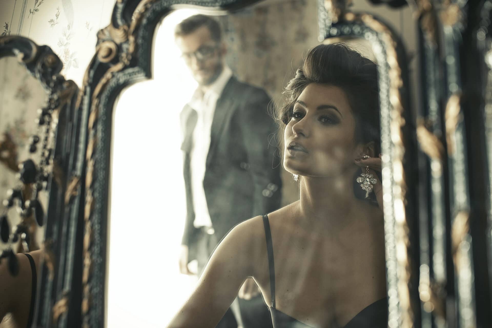 A woman looks into an ornate mirror, wearing a dark top and large earrings, pondering how to tell if you love someone. Her reflection is visible in the foreground, evoking thoughts about relationship dynamics, while a man in a suit stands slightly blurred in the background—symbolizing potential communication challenges. The room's elegant decor sets a contemplative scene for this simple love test, highlighting themes of self-reflection and potential marriage counselling or couples counselling to explore deeper emotional connections. Rosen Couples Counselling