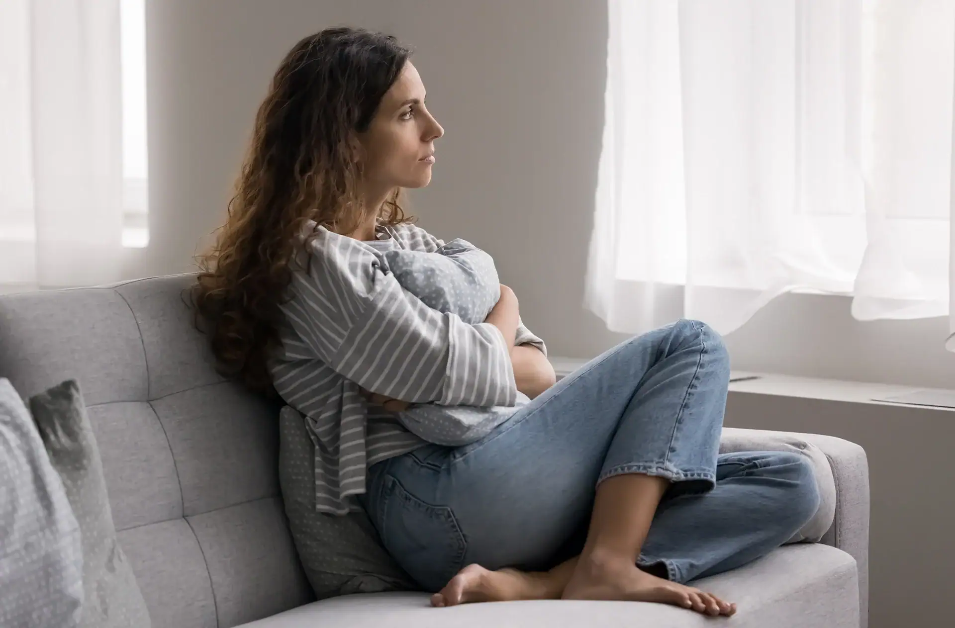 serious pensive young woman thinking on problem hugs cushion seated on couch at home