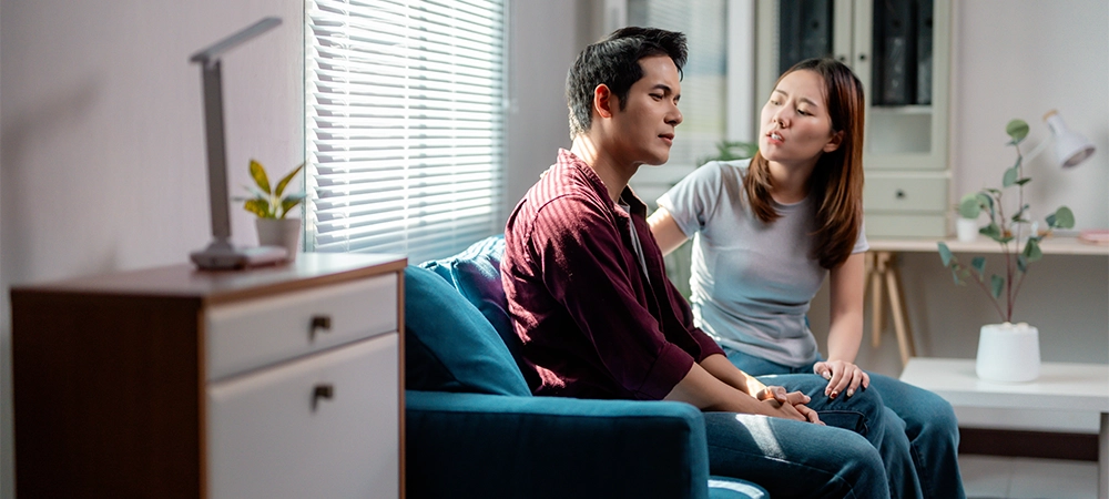 Young asian couple having relationship problems, woman trying to comfort her sad and stressed boyfriend sitting on the sofa at home